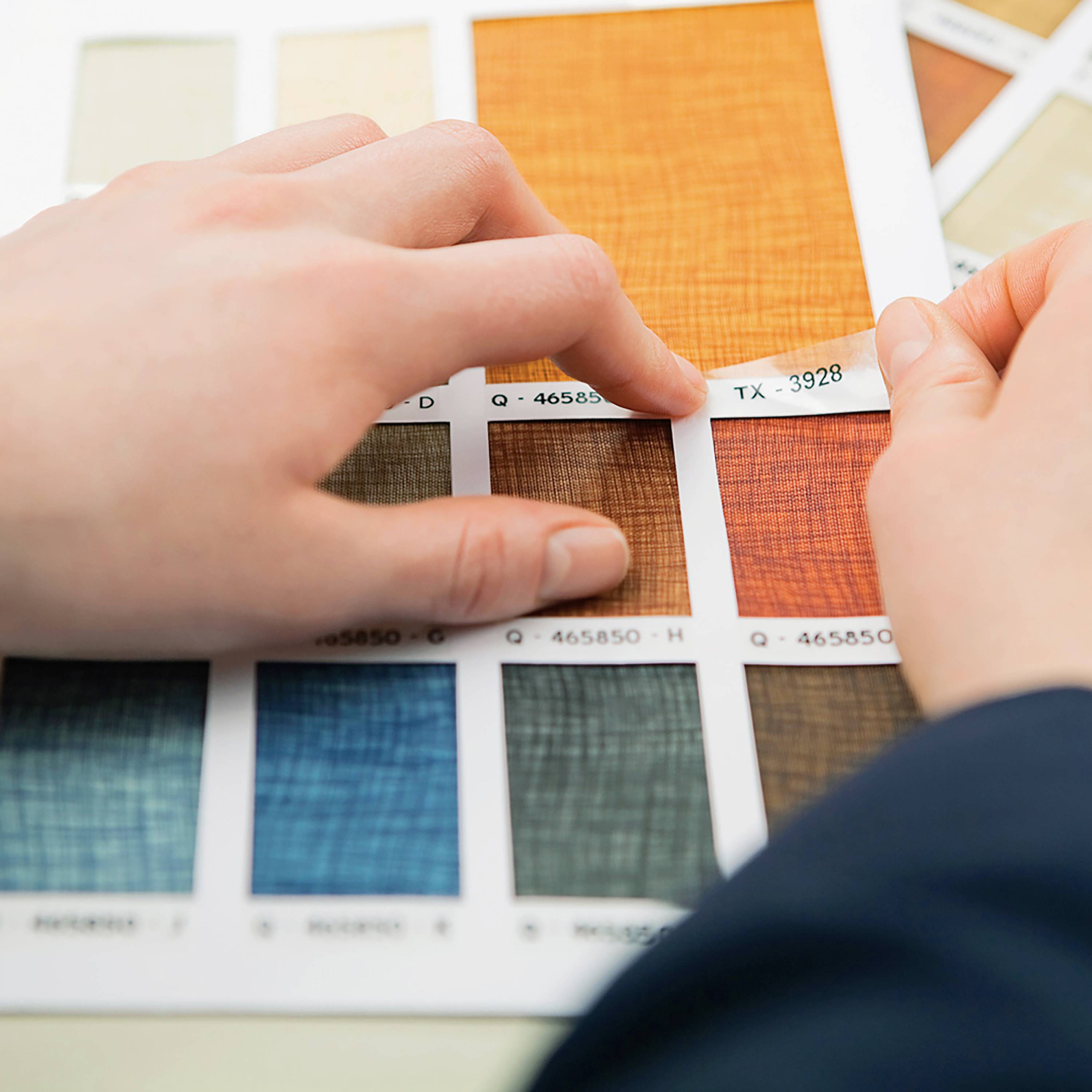 Une main tient un échantillon textile orange portant l'étiquette 'TX 3928' au-dessus d'une sélection d'autres motifs de tissus colorés.