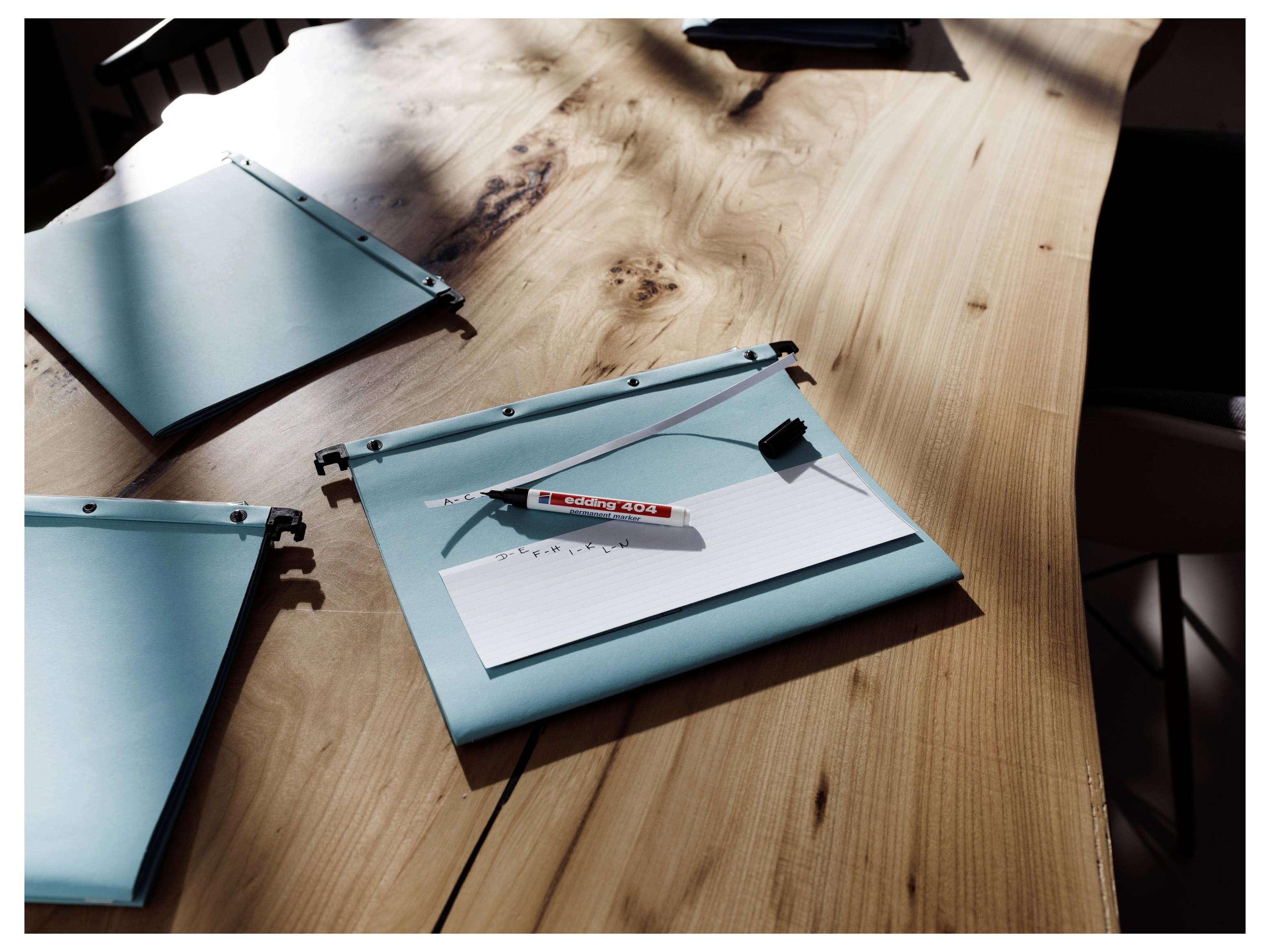 Trois dossiers bleus contenant des papiers et un stylo sont éparpillés sur un bureau en bois à la lumière du soleil éclatant.