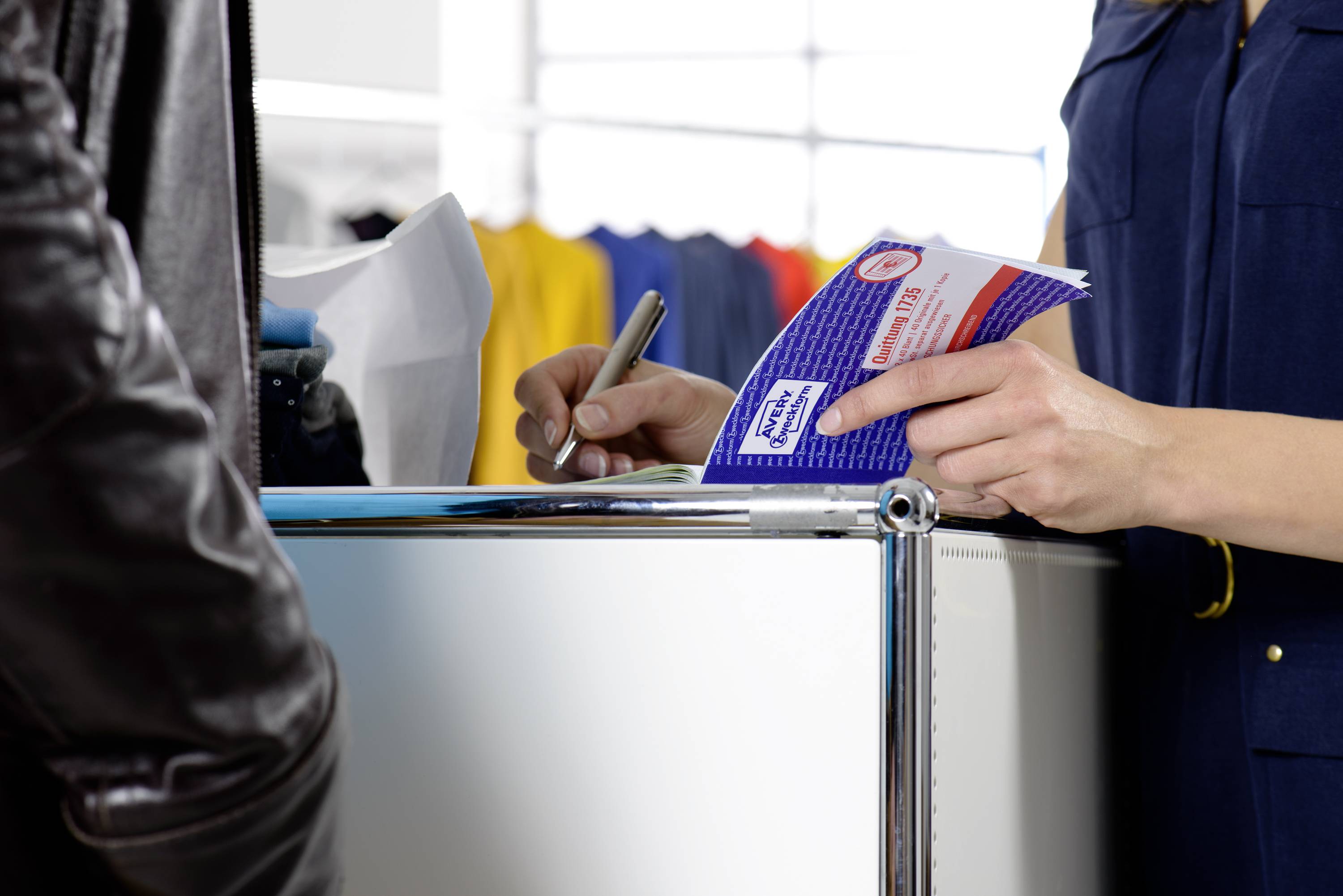 Une personne remet une brochure à une autre personne à un comptoir d'accueil. Des vêtements colorés sont visibles en arrière-plan.