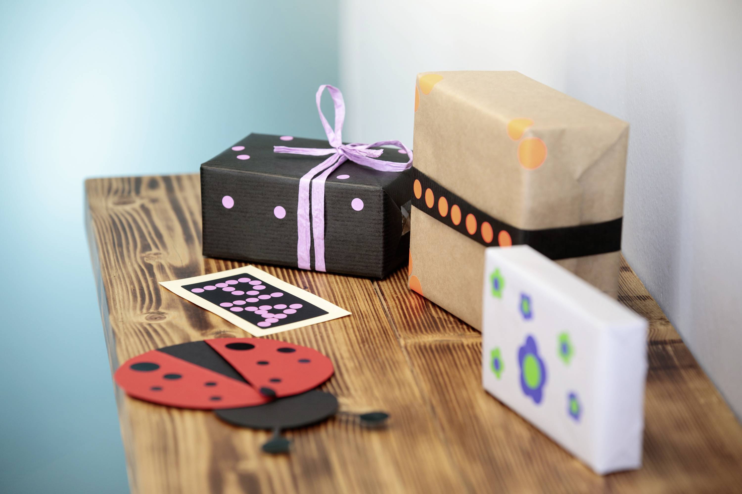Plusieurs cadeaux emballés de manière décorative sur une table en bois. L'un est orné d'un ruban violet, d'autres sont décorés de papier coloré et de points.