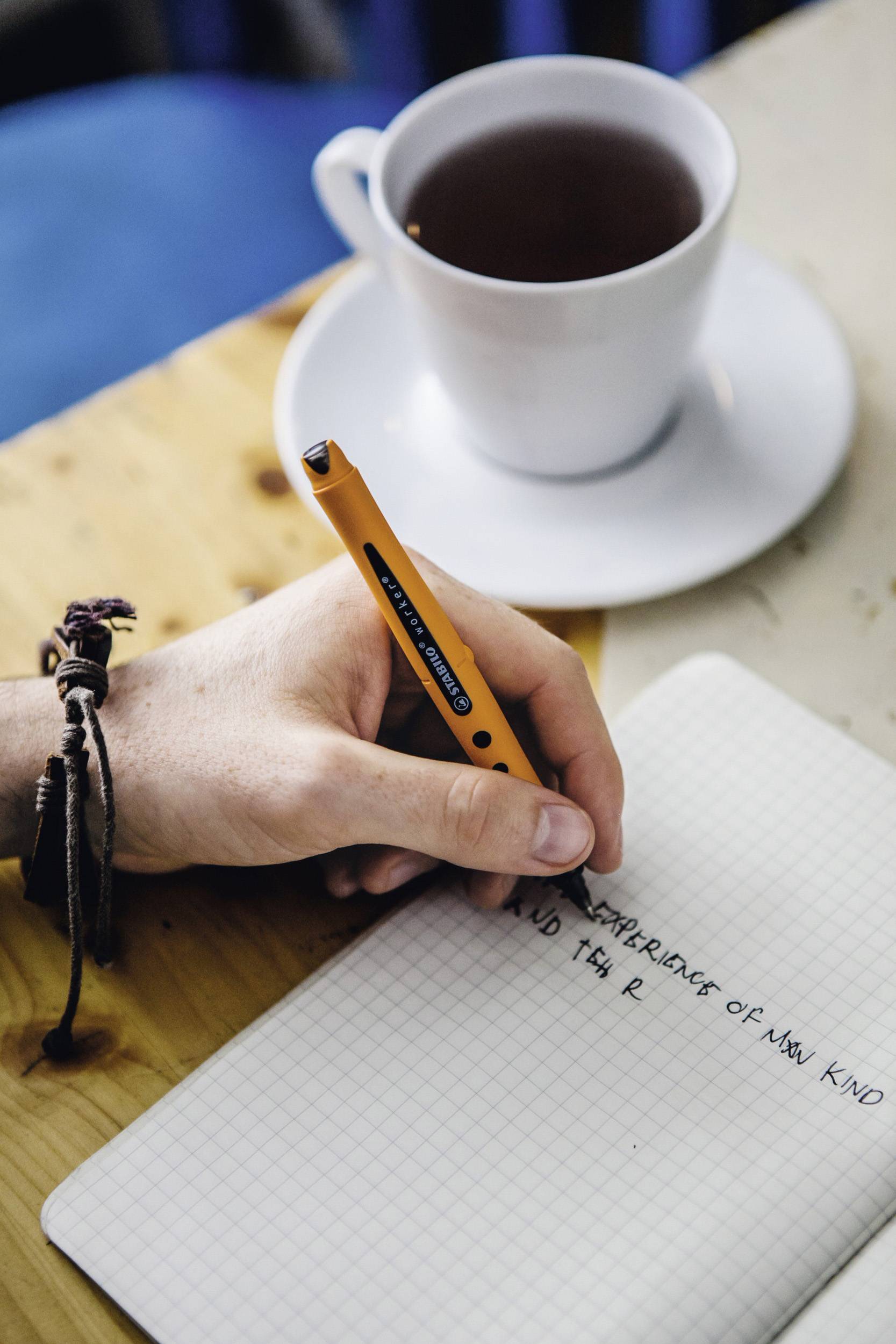 Une personne écrit dans un carnet posé sur une table. À côté se trouve une tasse de café.