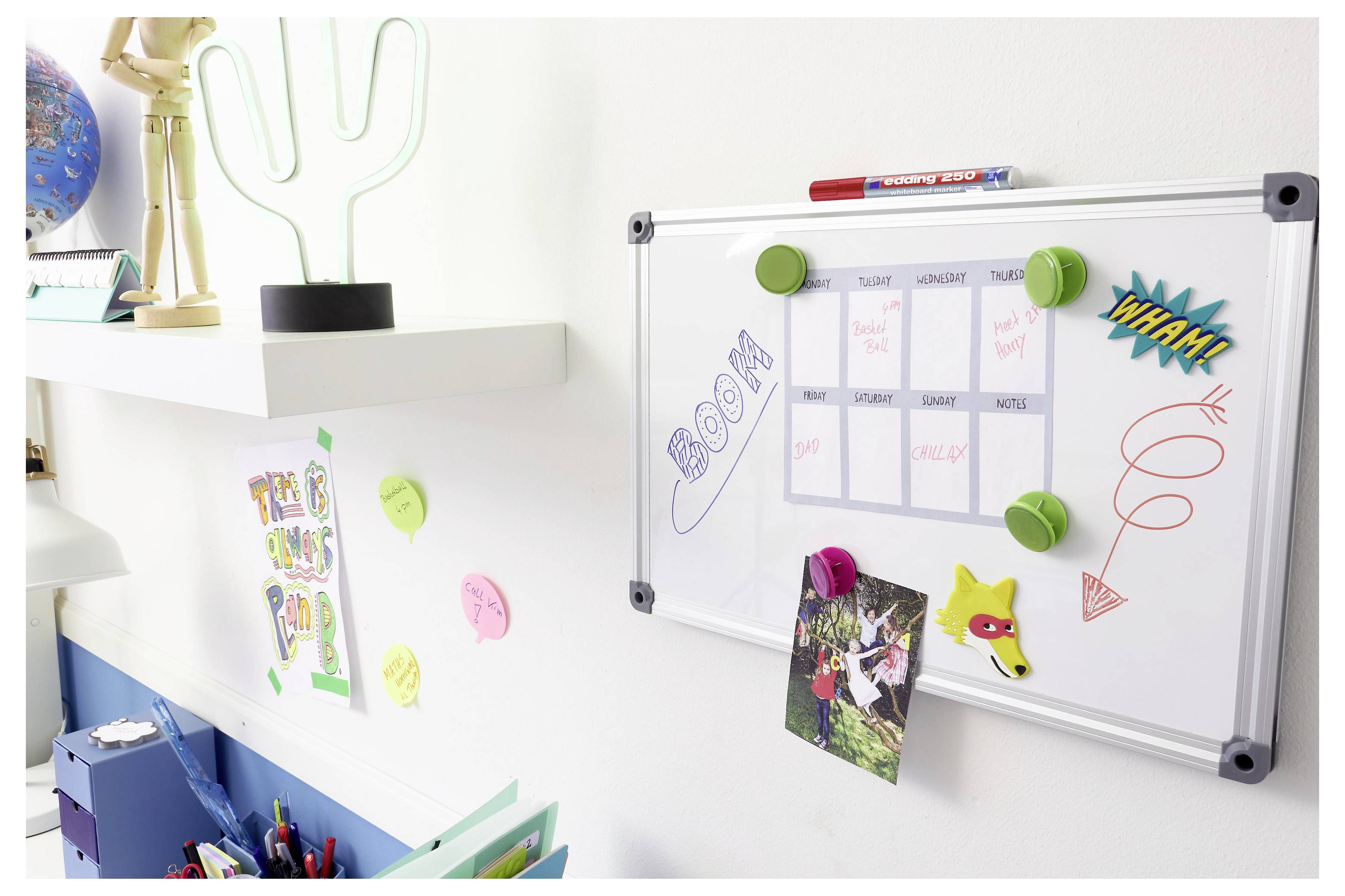Calendrier sur tableau blanc sur un mur avec des aimants colorés et des autocollants, à côté de dessins d'enfants et de fournitures de bureau dans un espace d'étude.