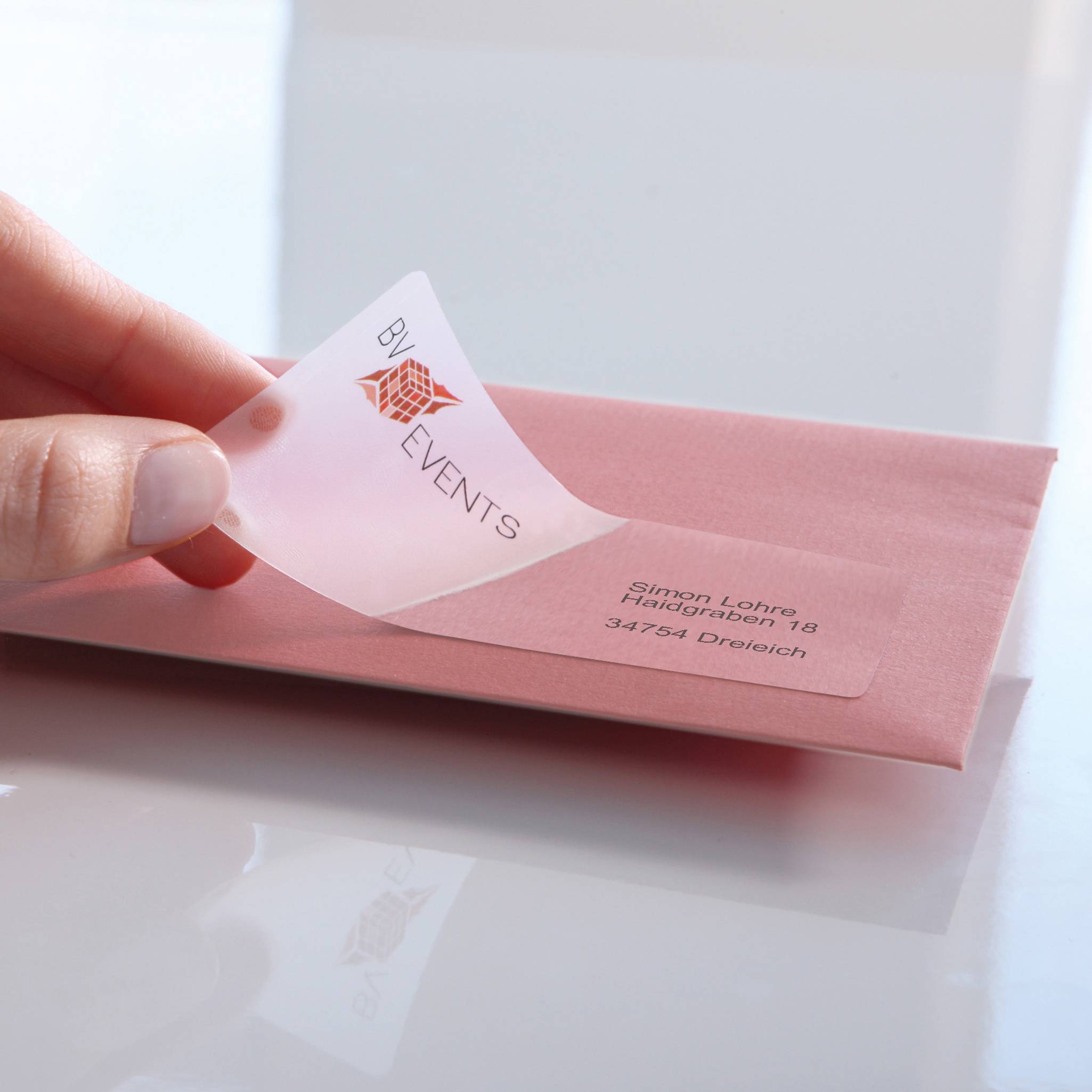 Une main colle une étiquette d'adresse transparente portant l'inscription 'BY EVENTS' sur une enveloppe rose.