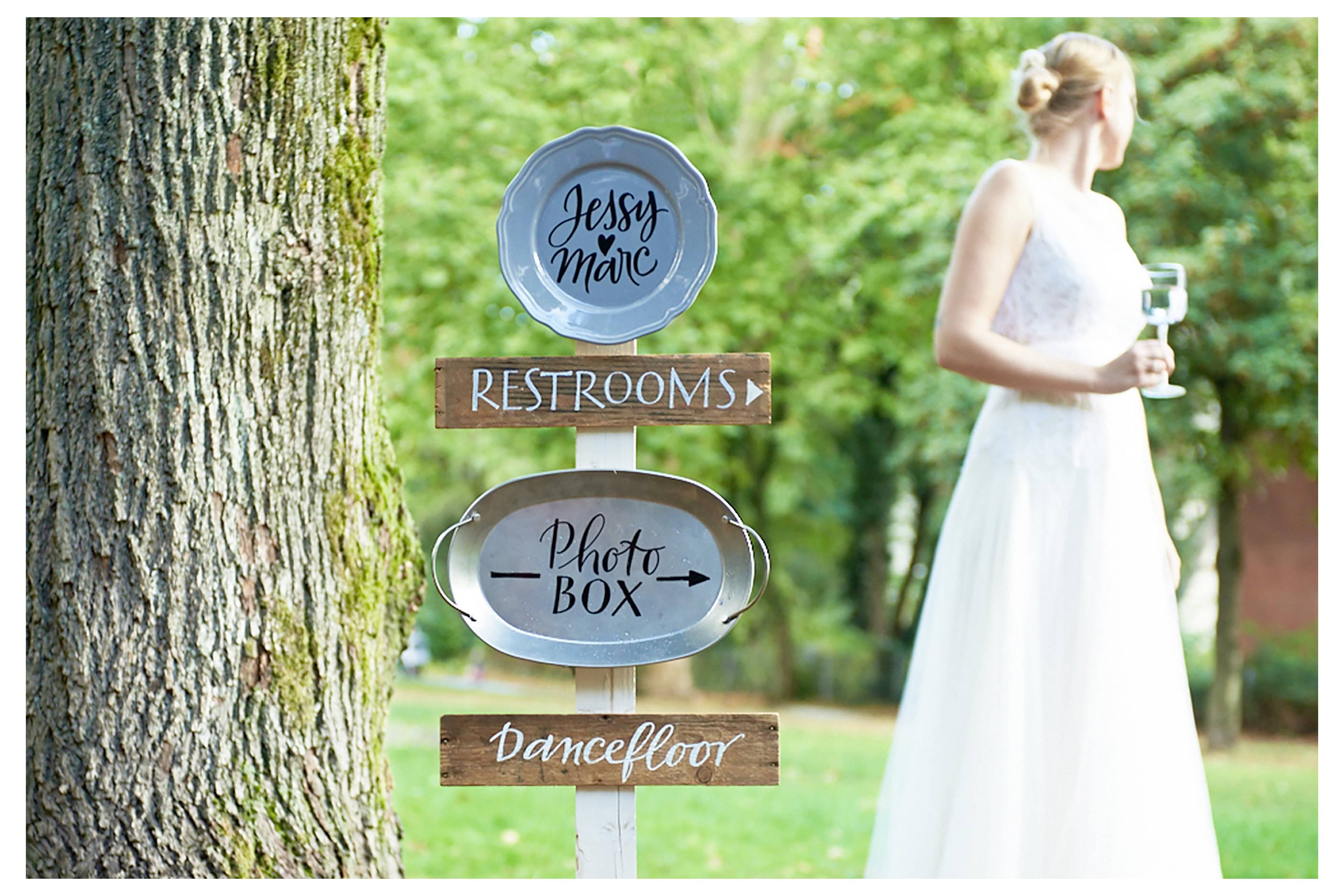 Panneau directionnel de mariage avec des flèches indiquant « TOILETTES », « Photobooth » et « Piste de danse » près d'une mariée en robe blanche en plein air.