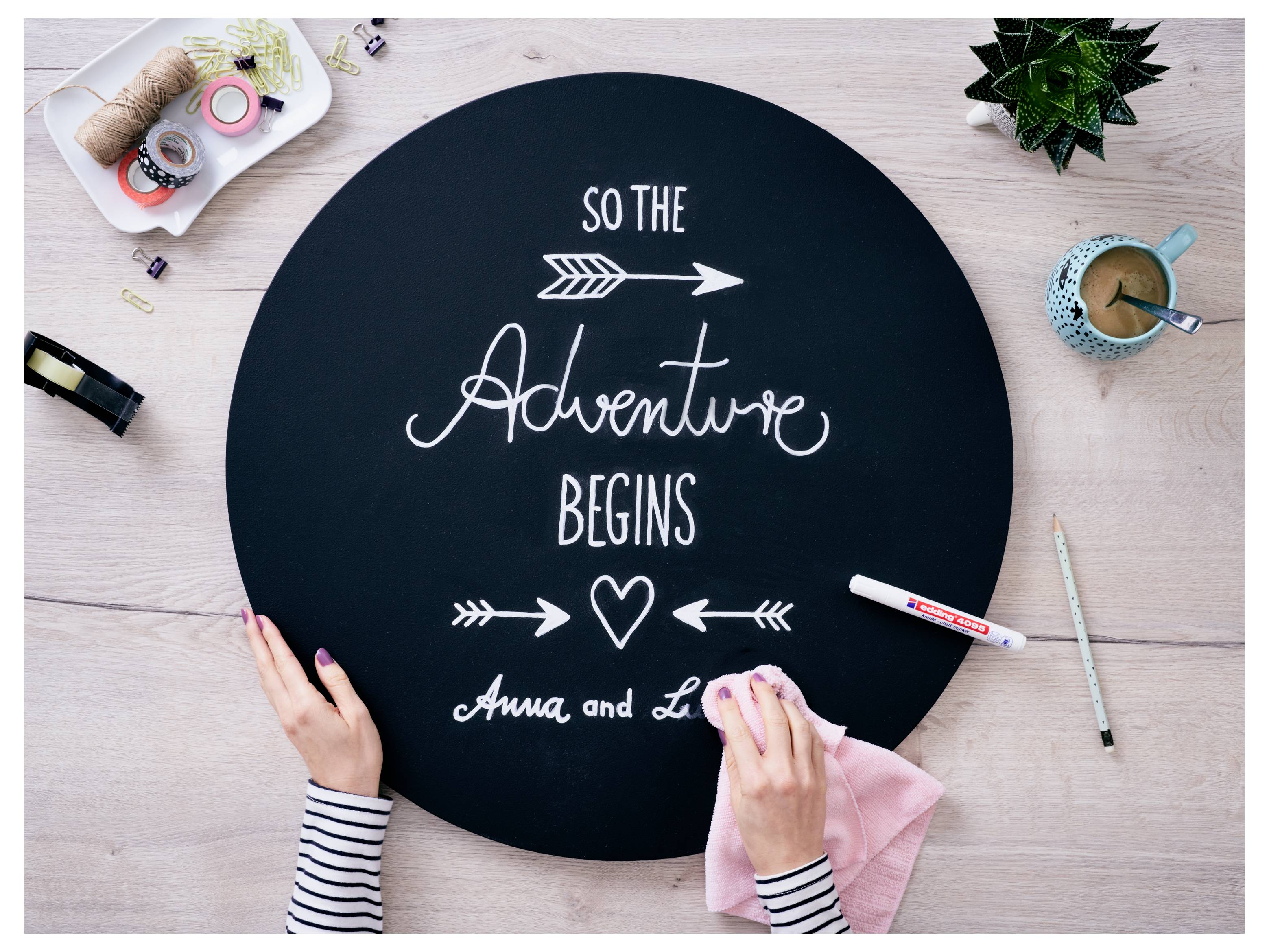 Des mains nettoient une pancarte ronde en ardoise noire lisant « Alors l'Aventure Commence » avec des flèches, un cœur et les noms « Anna et Lo » sur une table d'artisanat.