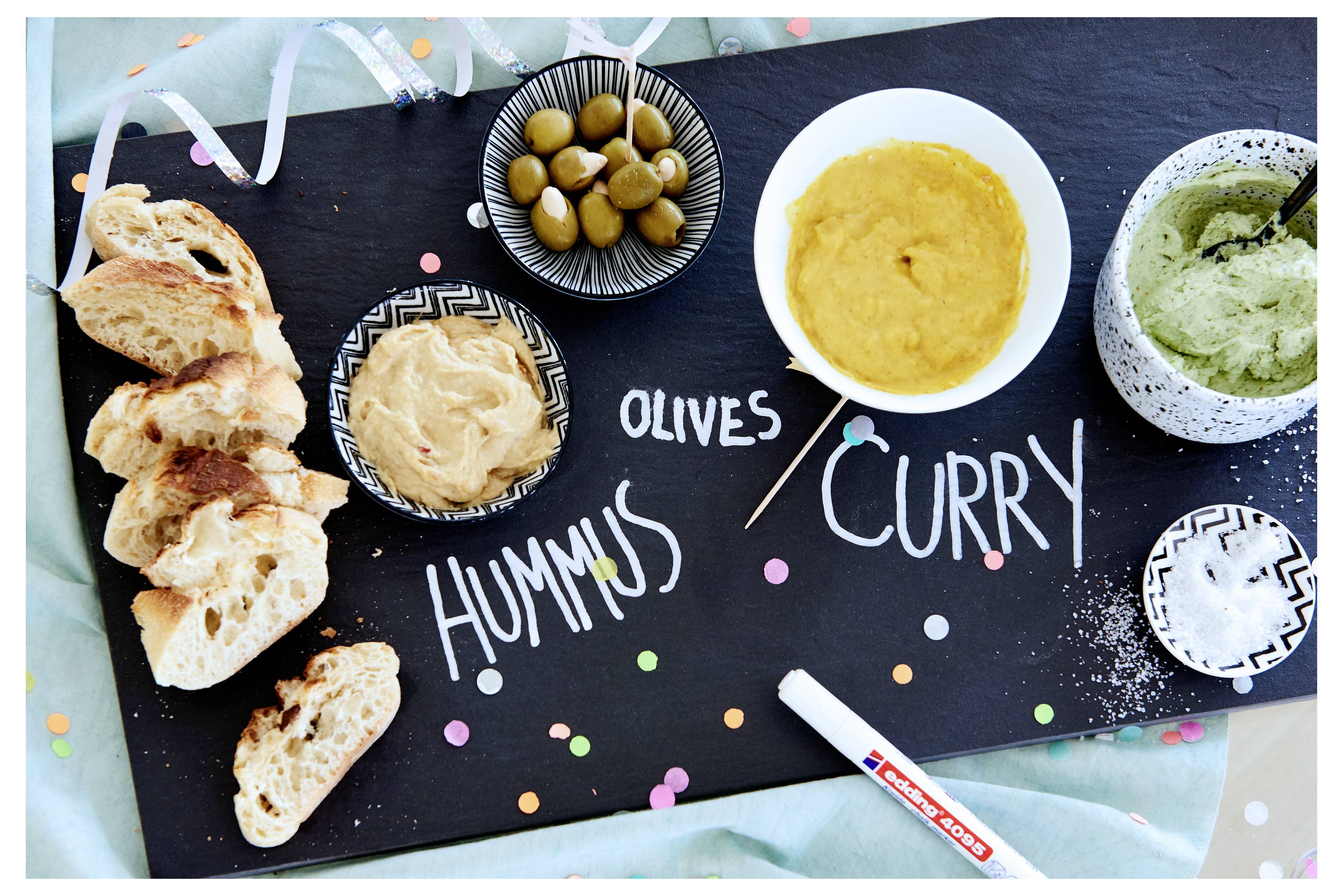 Assortiment de trempettes et de pain sur un tableau noir étiqueté « HUMMUS », « OLIVES » et « CURRY », avec des morceaux de pita et des bols d'olives, hummus, curry et trempette verte.