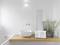 Une salle de bain minimaliste avec un lavabo blanc sur une table en bois, à côté duquel se trouvent un petit poste de radio et une plante.