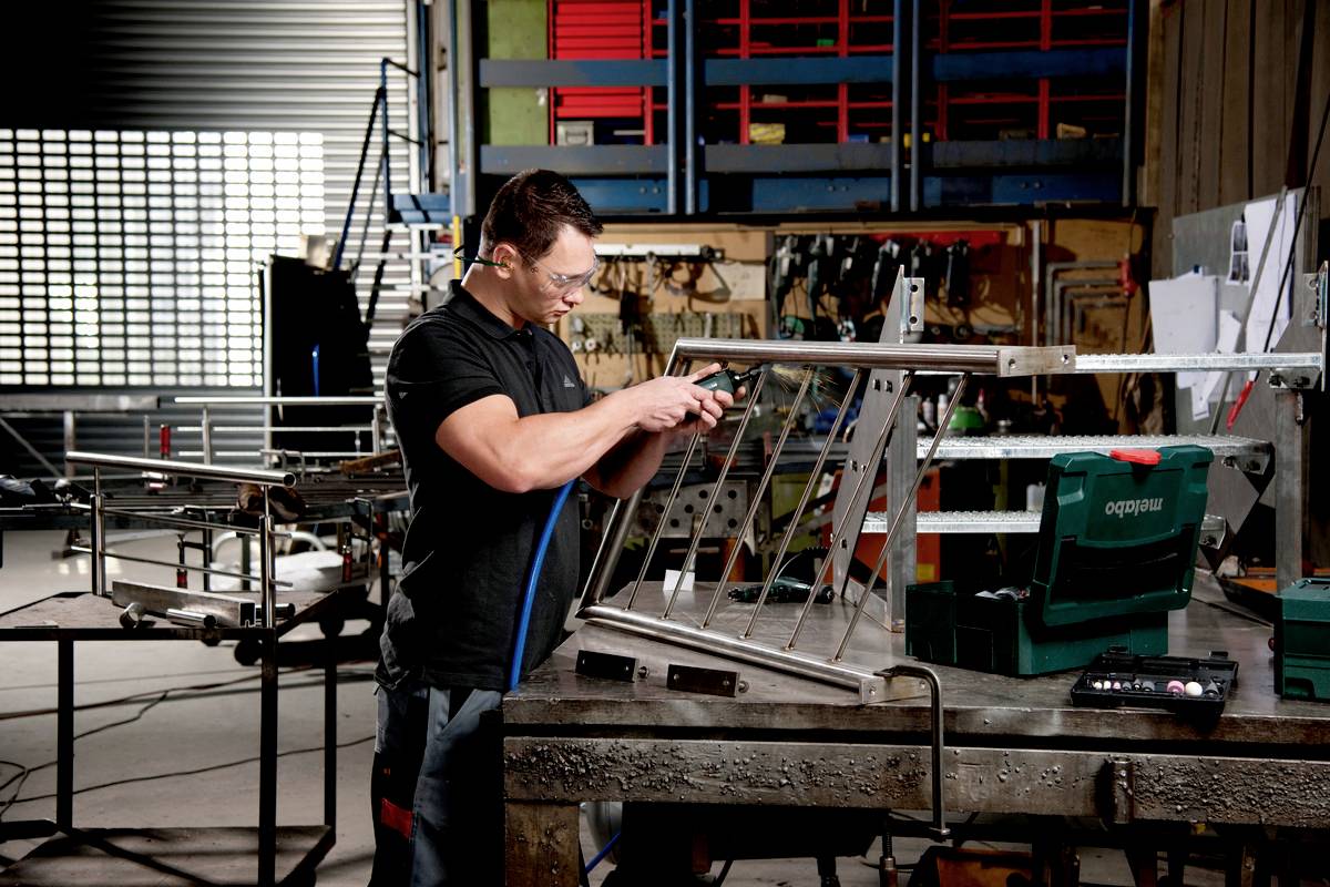 Un homme dans un atelier répare un châssis métallique à l'aide d'outils. Des étagères et des équipements sont visibles en arrière-plan.