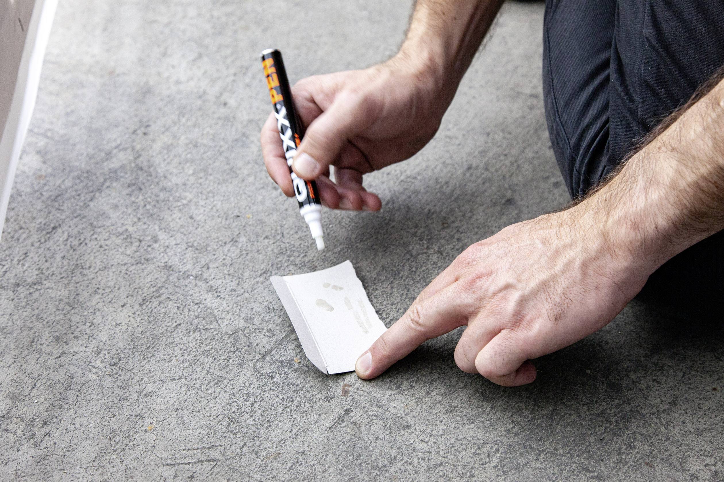 Un homme écrit avec un stylo blanc sur une petite feuille de papier posée sur un sol en béton gris.