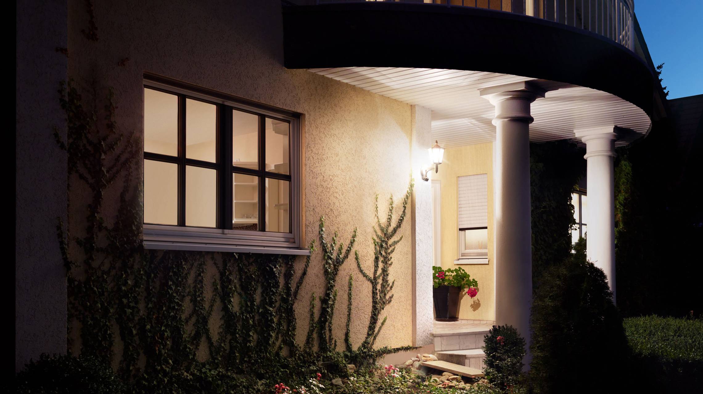 Une maison avec une entrée illuminée la nuit, entourée de plantes. Deux colonnes soutiennent l'avant-toit.