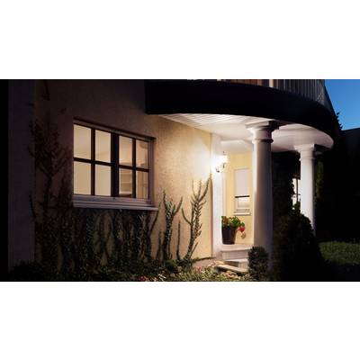 Une maison avec une entrée illuminée la nuit, entourée de plantes. Deux colonnes soutiennent l'avant-toit.
