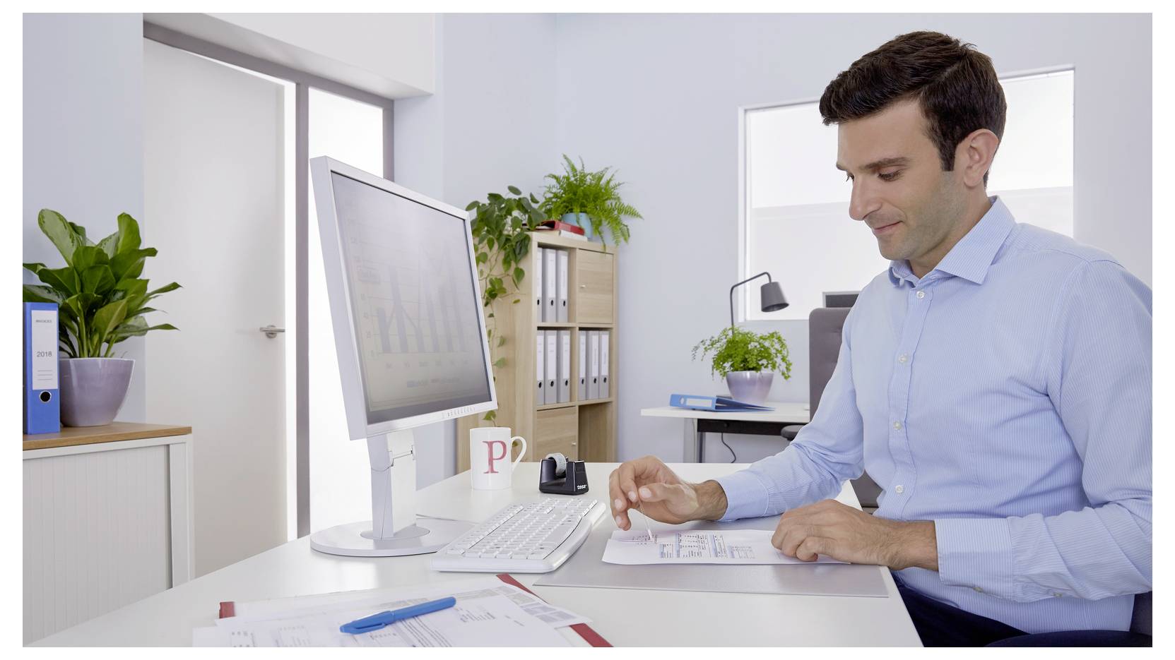 Un homme en chemise bleue est assis à un bureau dans un bureau moderne, examinant des documents. Un écran d'ordinateur affiche un graphique. Des plantes et des dossiers sont visibles.