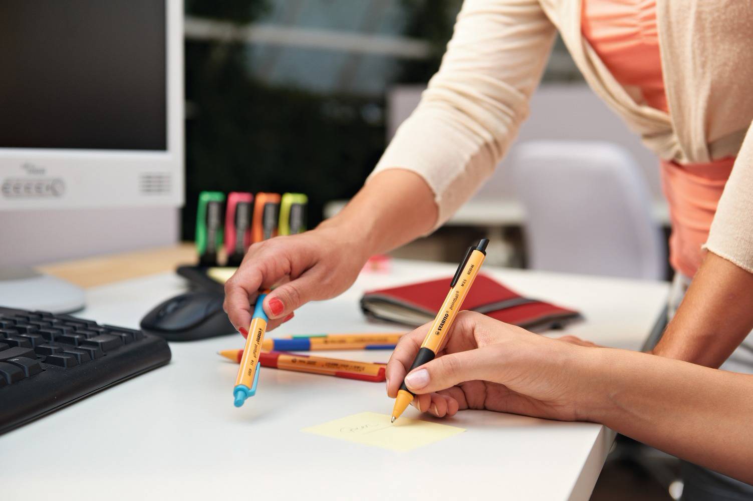 Deux personnes travaillent à un bureau ; l'une pointe une note sur un post-it. Divers stylos et fournitures de bureau sont visibles.