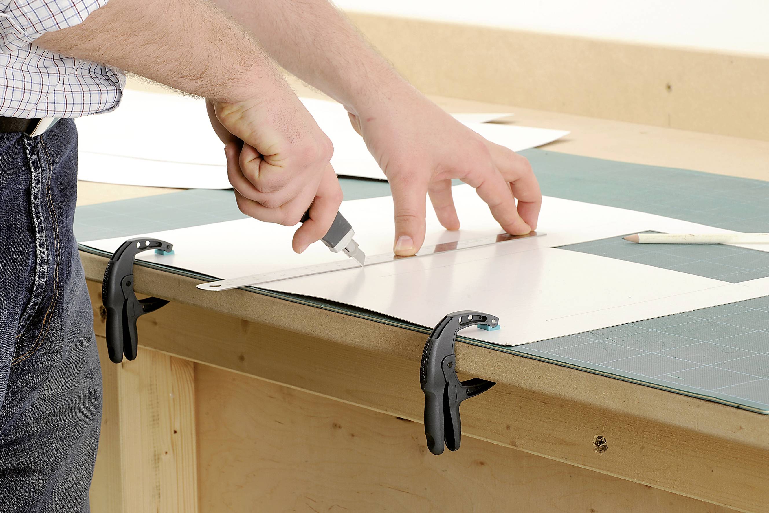 Une personne découpe précisément un carton blanc avec un cutter, fixé sur une table de travail à l'aide de pinces.