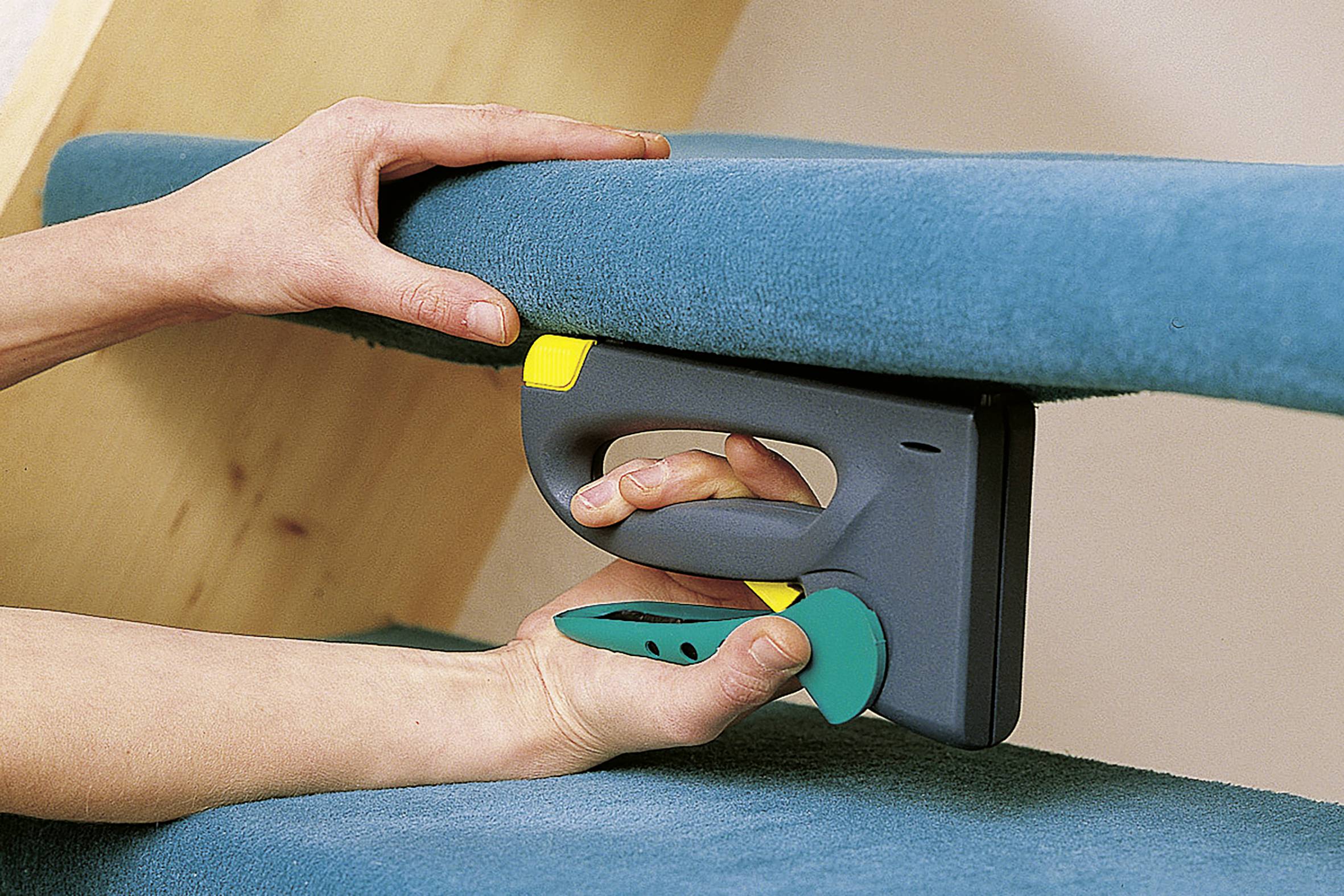 Une personne utilise un pistolet à agrafes électrique pour fixer du tissu bleu sur une surface en bois. Les mains tiennent le tissu tendu.