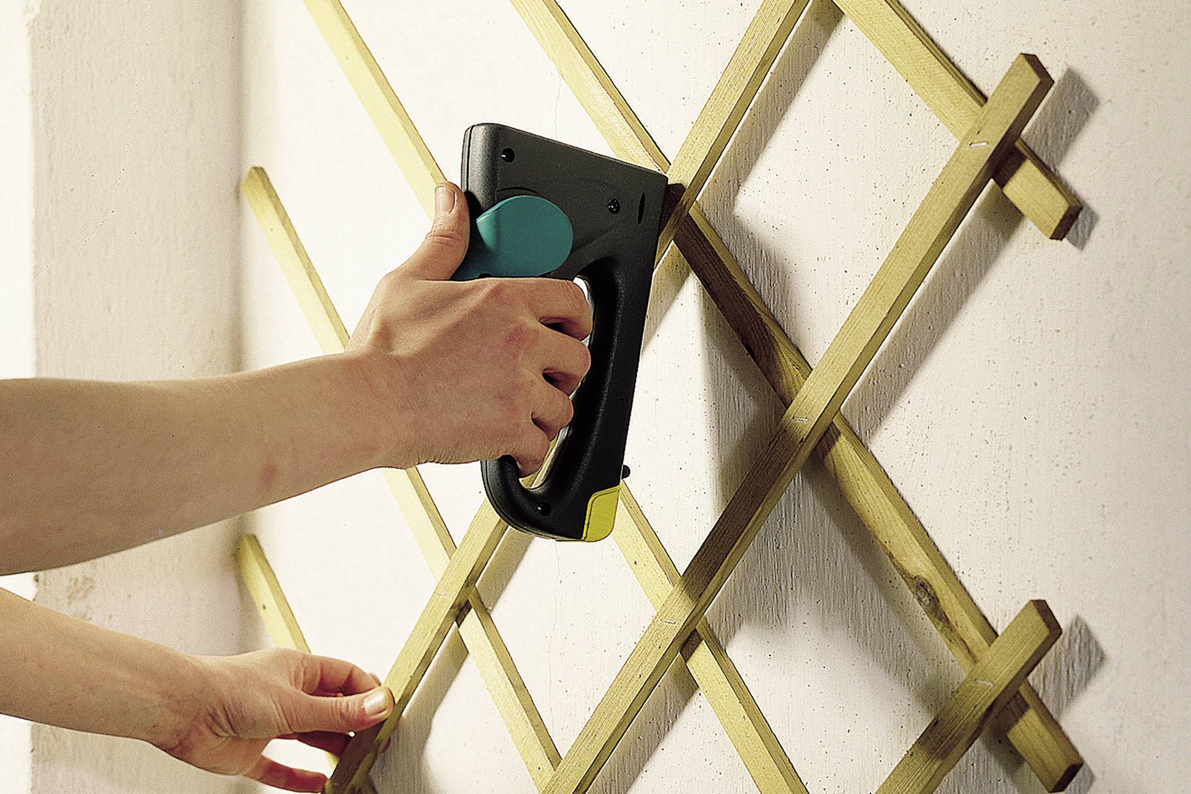 Une main utilise un pistolet à agrafes pour fixer une grille en bois sur un mur.