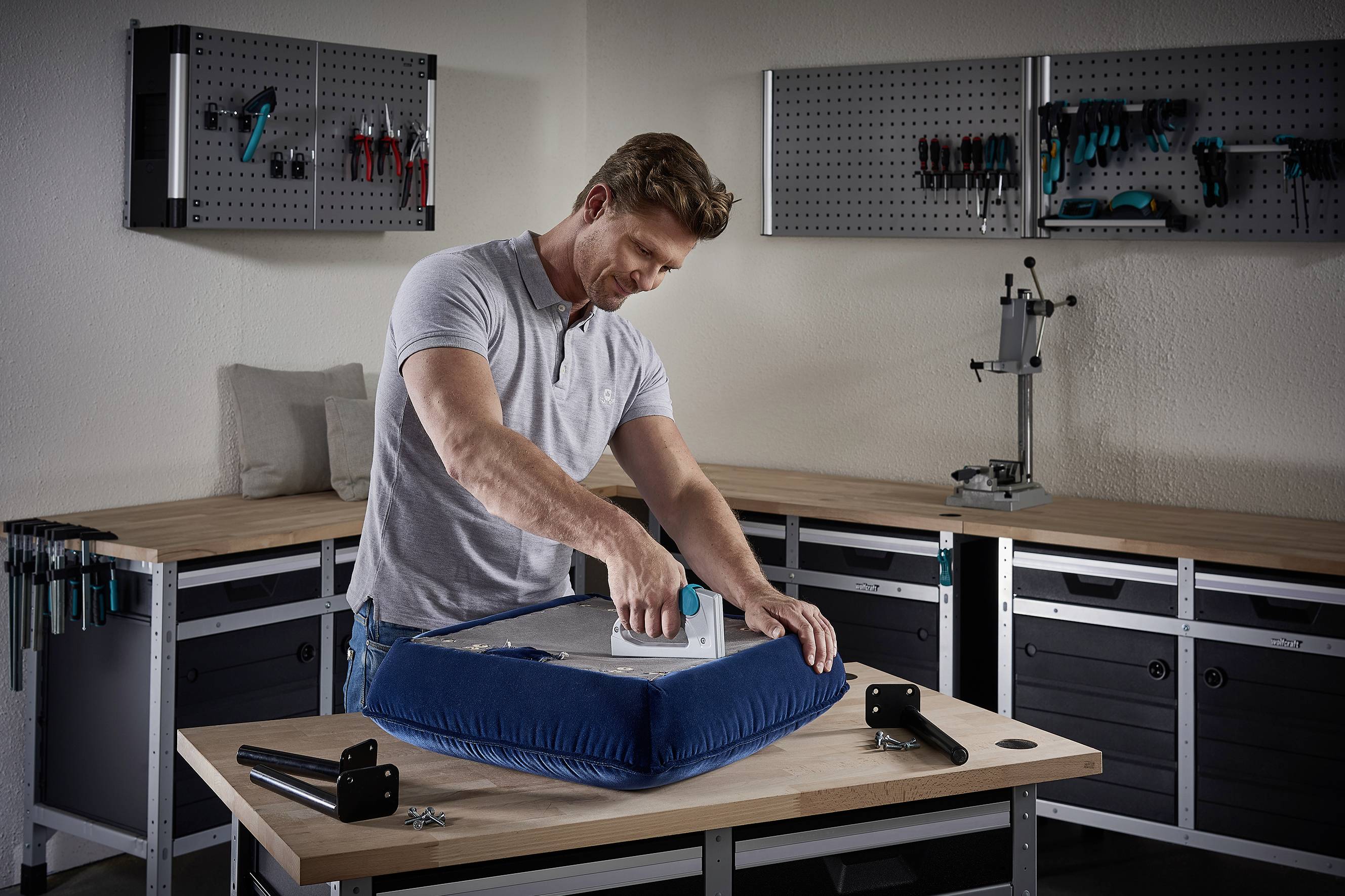 Un homme recouvre un siège d'un revêtement en tissu bleu dans un atelier. Des vis et des pieds de chaise sont disposés sur la table.