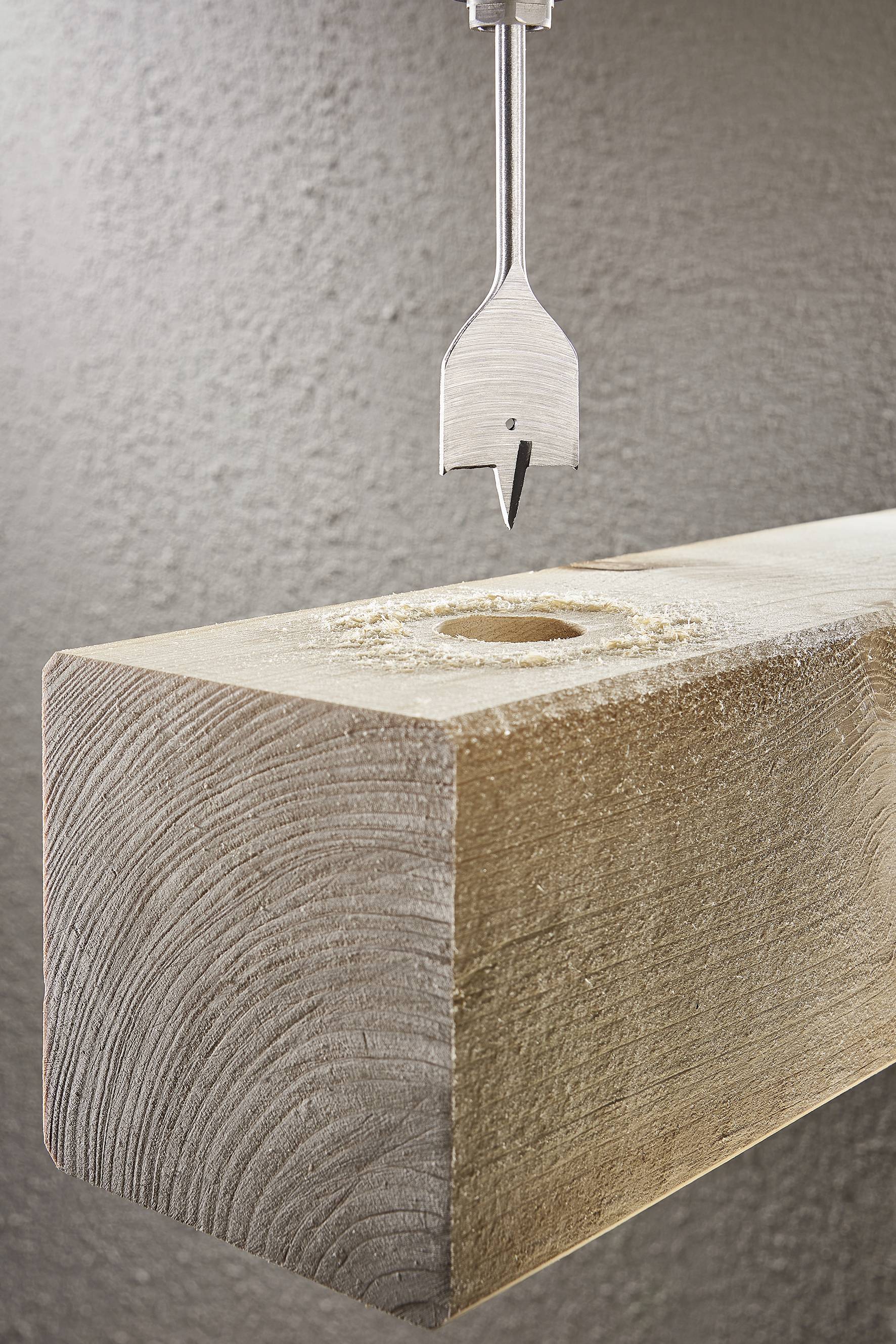 Un foret hélicoïdal plane au-dessus d'une poutre en bois avec un trou percé. L'arrière-plan est structuré.