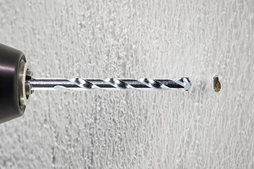 Un foret perce un trou dans un mur blanc et structuré. La pointe du foret est argentée et pénètre proprement dans le mur.