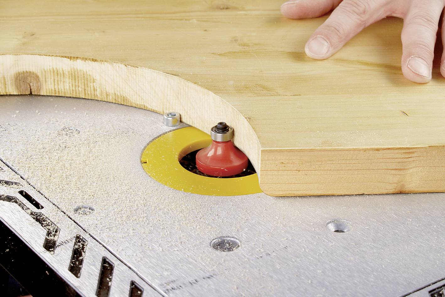 Une main guide une planche en bois sur une fraiseuse pour créer un bord arrondi. Des copeaux de bois sont visibles.