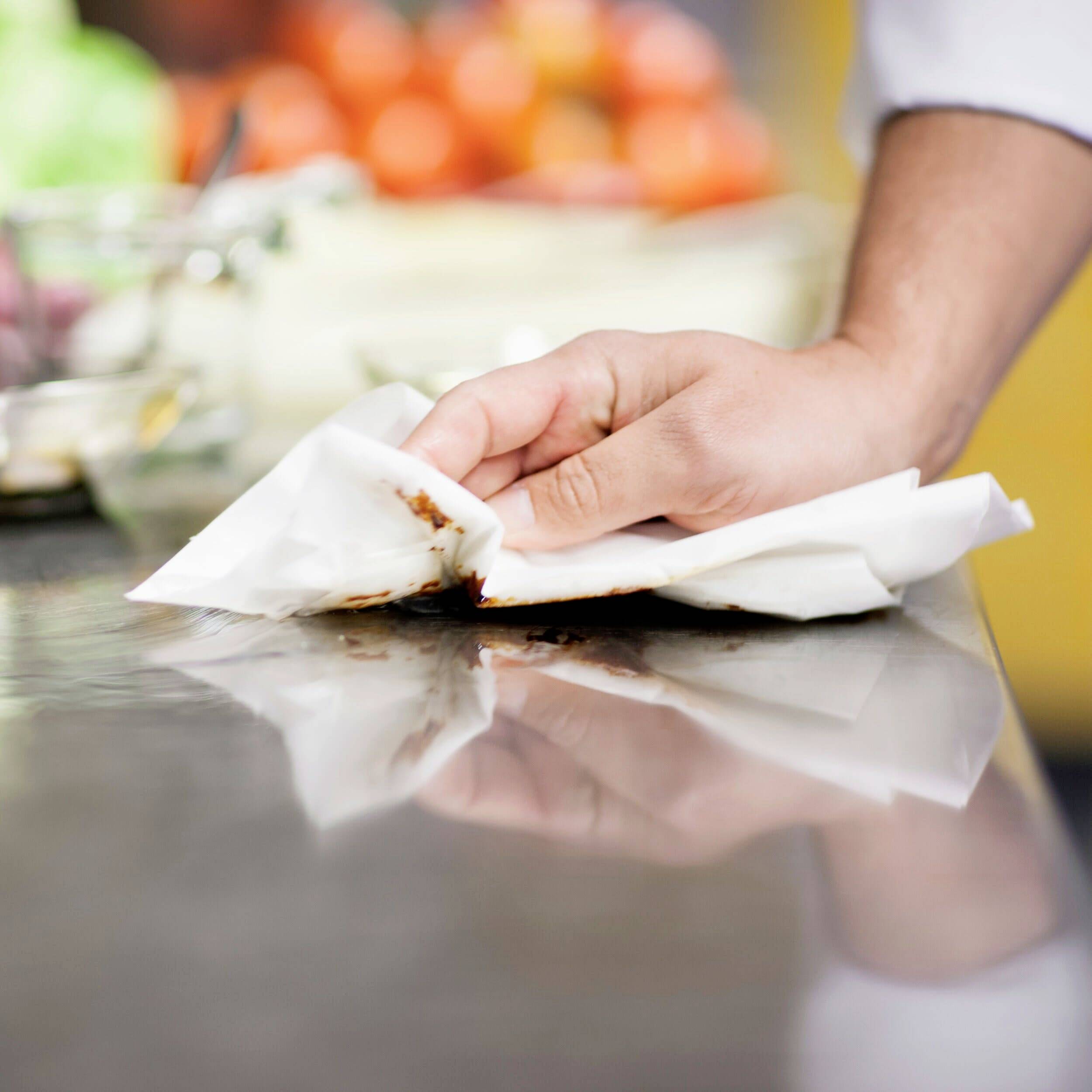 Une main nettoie une surface sale avec un essuie-tout dans une cuisine. Des ingrédients flous sont visibles en arrière-plan.