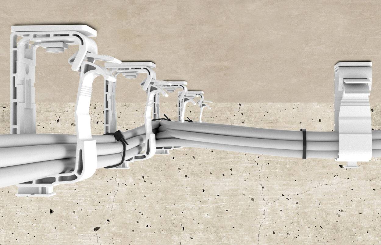 Plusieurs supports de câbles blancs fixés à un mur en béton maintiennent des câbles regroupés. Les supports sont disposés à intervalles réguliers.