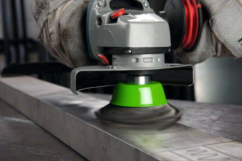 Une main tient une meuleuse d'angle qui rectifie du métal avec un disque de meulage vert. Des étincelles volent, des gants de travail protègent la main.