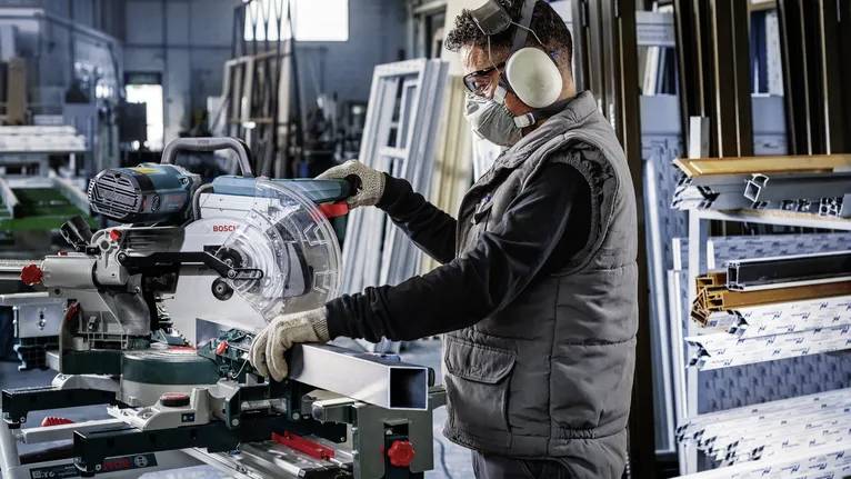Une personne portant un équipement de sécurité utilise une scie électrique dans un atelier avec des structures métalliques et des outils en arrière-plan.