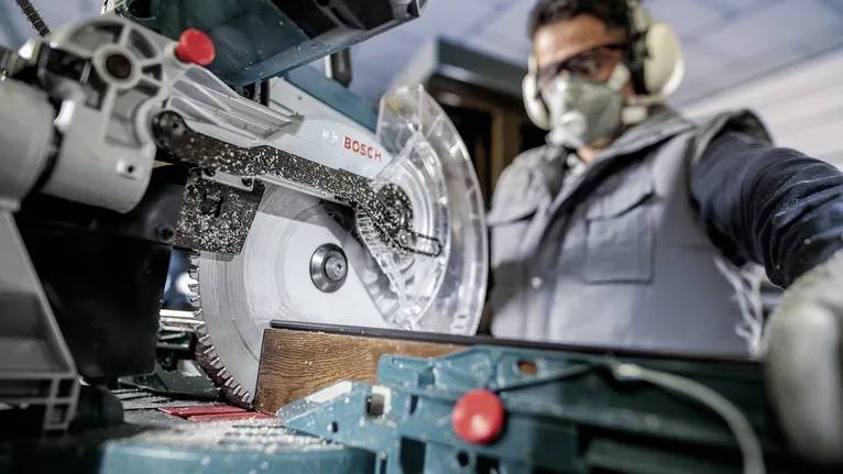 Un ouvrier utilise une scie électrique, découpant un morceau de bois. L'ouvrier porte un équipement de protection, comprenant des protections auditives et un masque.