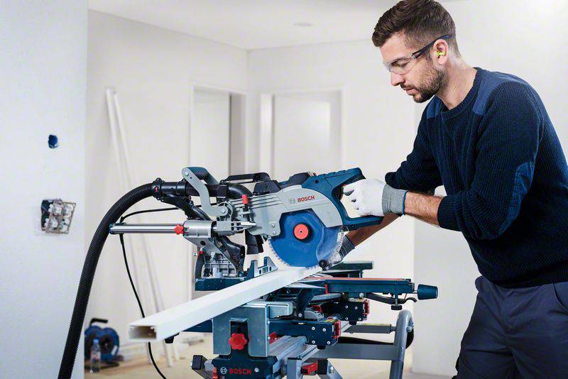 Un homme utilise une scie circulaire de table dans une pièce rénovée. Il porte des lunettes de protection et des gants. Des murs blancs sont visibles en arrière-plan.