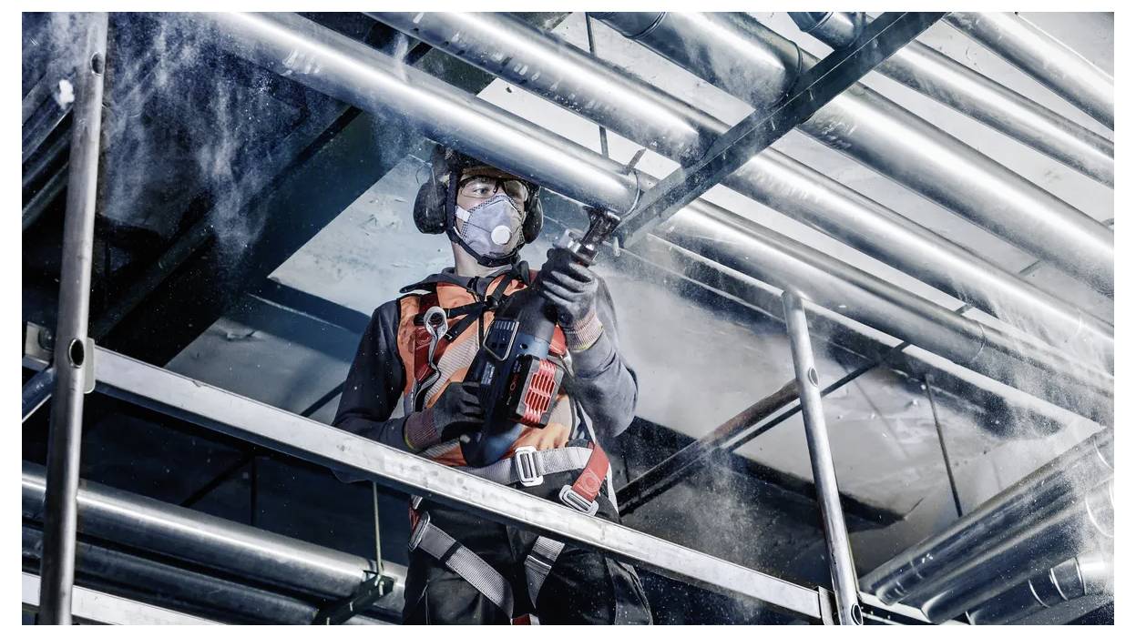 Travailleur en équipement de protection utilisant un tuyau haute pression sur des échafaudages industriels, entouré de tuyaux métalliques. Équipement de sécurité visible.