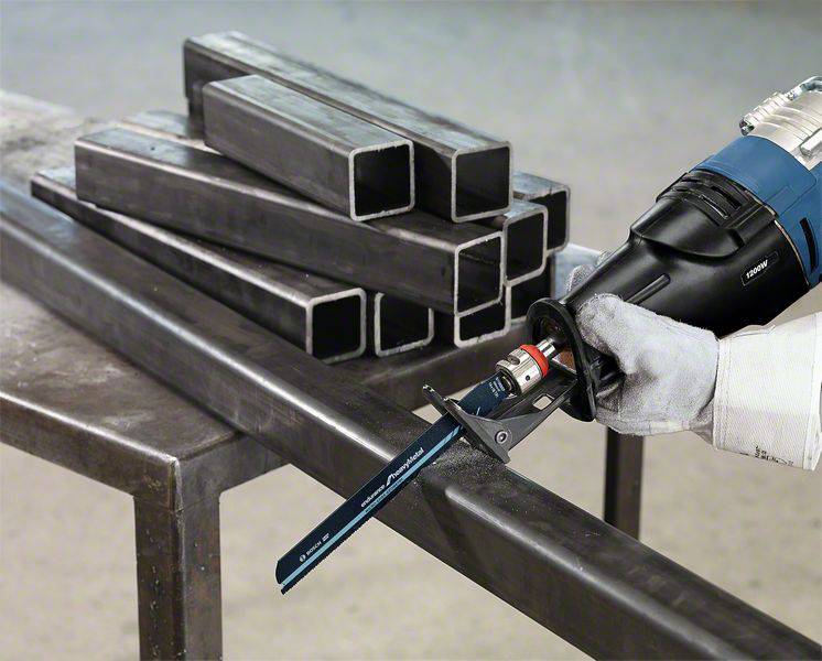Un ouvrier coupe un tube d'acier rectangulaire avec une scie électrique bleue sur une table. D'autres tubes sont posés en arrière-plan.