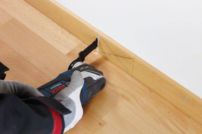 Une personne coupe un plancher en bois avec une scie électrique à côté d'une plinthe. Elle porte des gants de travail.
