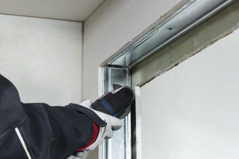 Une personne vêtue de vêtements de travail coupe un renfort métallique dans un chambranle de porte à l'aide d'un outil oscillant.