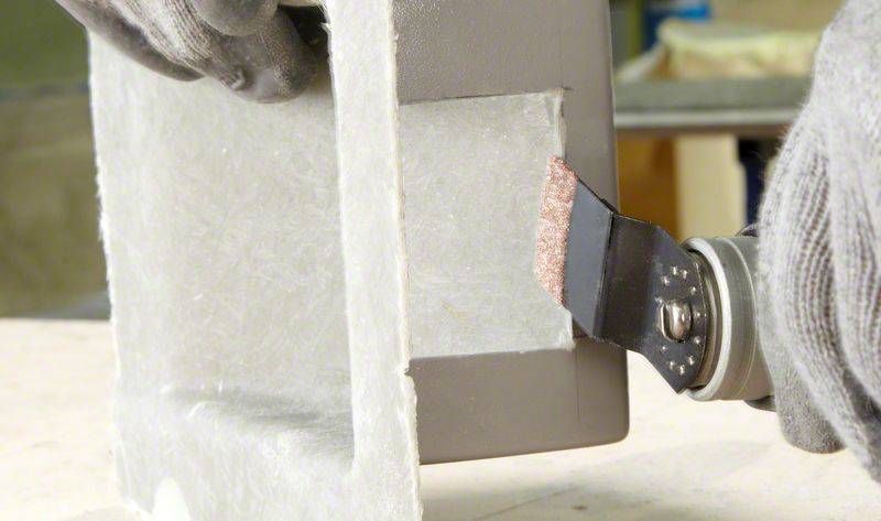 Une personne coupe un matériau gris à l'aide d'un outil multifonction oscillant. Porte des gants de protection.