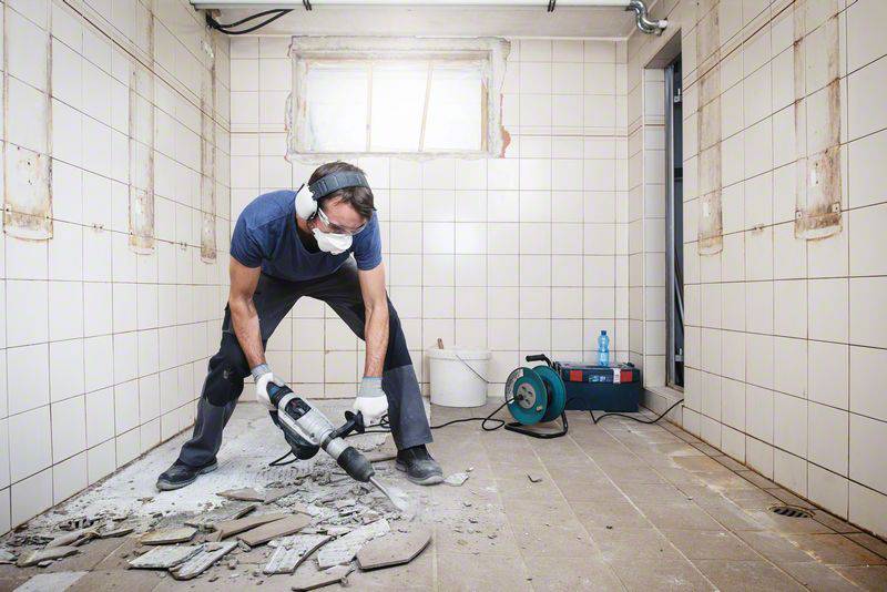 Une personne portant un équipement de protection utilise un marteau-piqueur pour enlever des carreaux dans une pièce carrelée vide. Environnement de chantier.