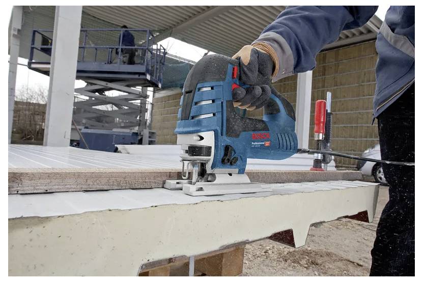 Une personne portant des gants utilise une scie sauteuse pour couper une planche de bois à l'extérieur. Un élévateur à ciseaux et un autre ouvrier sont visibles en arrière-plan.