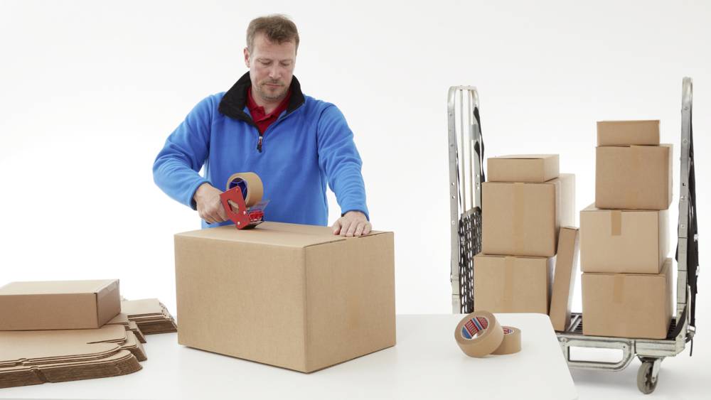 Un homme portant un pull bleu est en train de sceller un grand carton avec du ruban adhésif. À l'arrière-plan, d'autres cartons sont disposés sur un chariot roulant.