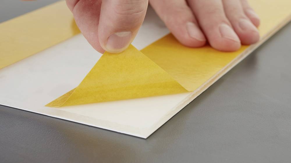 Une main retire du papier de protection jaune d'une surface adhésive sur une table grise.