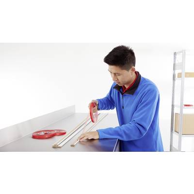 Un homme portant un pull bleu utilise un ruban adhésif rouge pour fixer des lattes de bois sur une table. Des étagères sont visibles en arrière-plan.