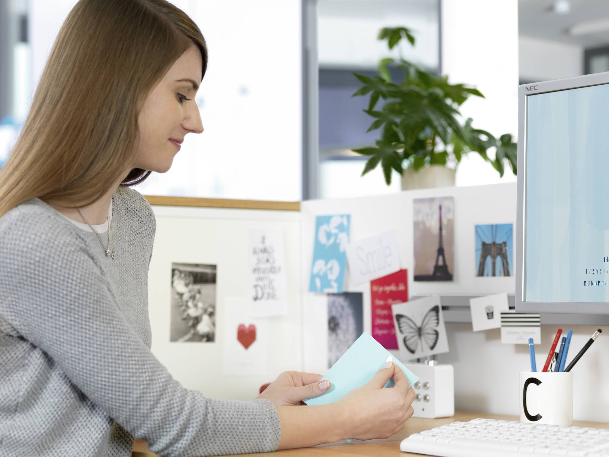 Une femme est assise à un bureau et lit un document. En arrière-plan, on peut voir des photos, des cartes et une plante. Bureau avec un écran d'ordinateur et une tasse.