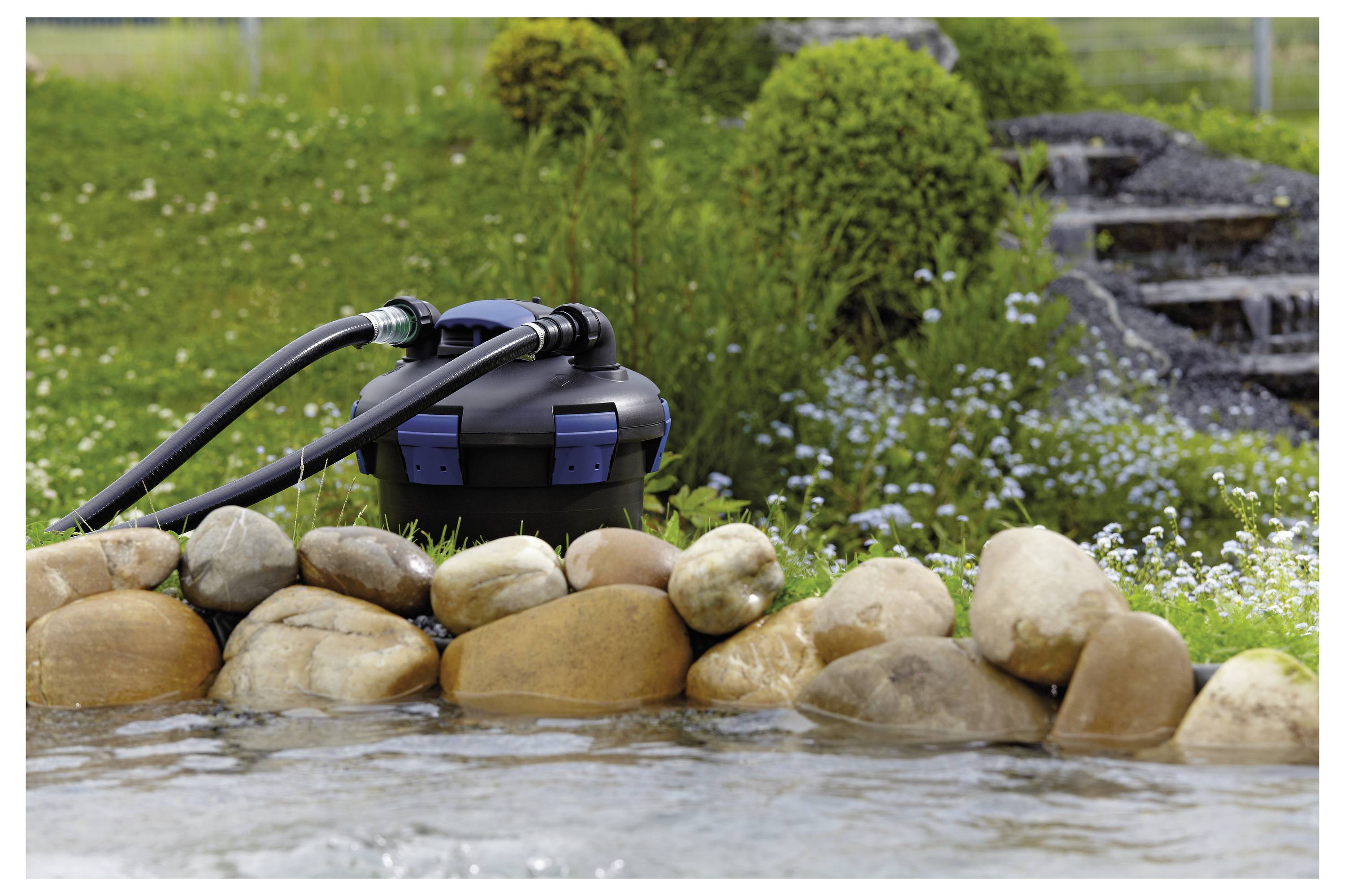 Filtre de bassin avec des tuyaux à côté d'un bassin bordé de pierres, entouré d'herbe et de fleurs, avec une petite cascade en arrière-plan.