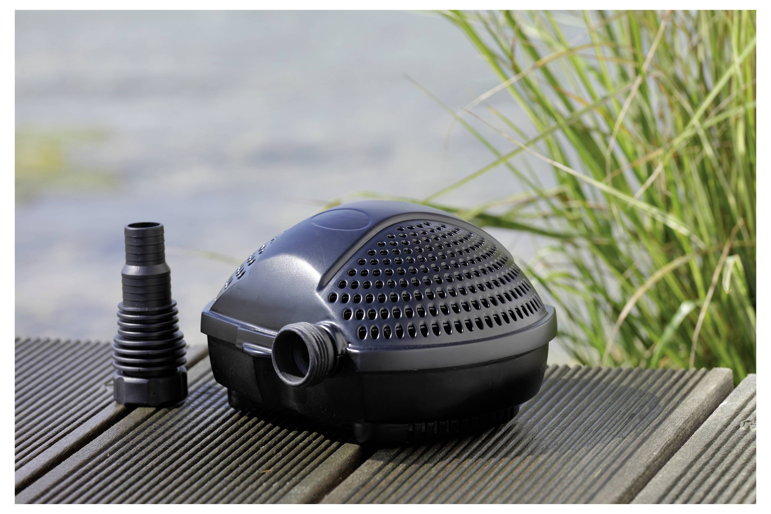 Une pompe de bassin noire est placée sur une terrasse en bois près d'une herbe haute et verte, avec un accessoire de déversement à côté. Un arrière-plan aquatique flou est visible.