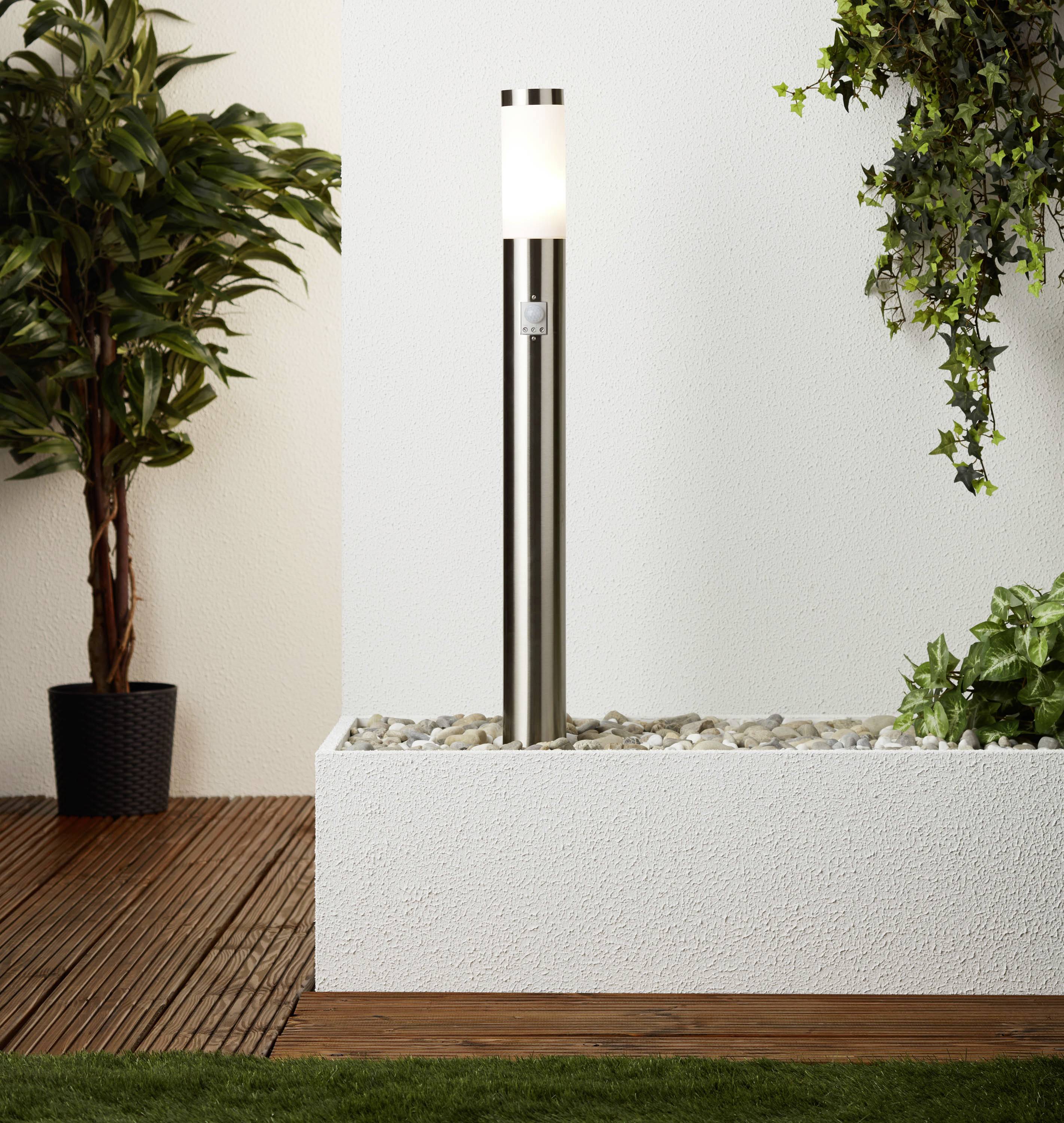 Un luminaire extérieur moderne en acier inoxydable est posé sur un lit de graviers, entouré de plantes. Il éclaire une terrasse avec des détails de bois et d'herbe.