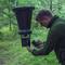 Un homme dans un environnement extérieur installe un distributeur suspendu, versant de la nourriture à l'intérieur. Le distributeur est accroché à des arbres, entouré de verdure.