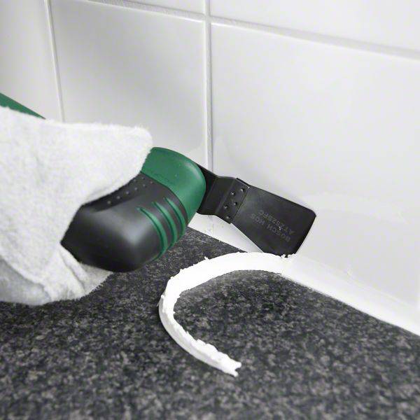 Quelqu'un utilise un outil pour enlever le mastic d'étanchéité entre les carreaux et le plan de travail dans une cuisine.