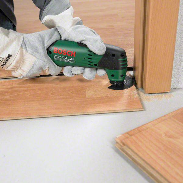 Une personne coupe un plancher en bois au niveau d'un seuil de porte à l'aide d'un outil multifonction Bosch vert. Les mains sont gantées de gants de travail.