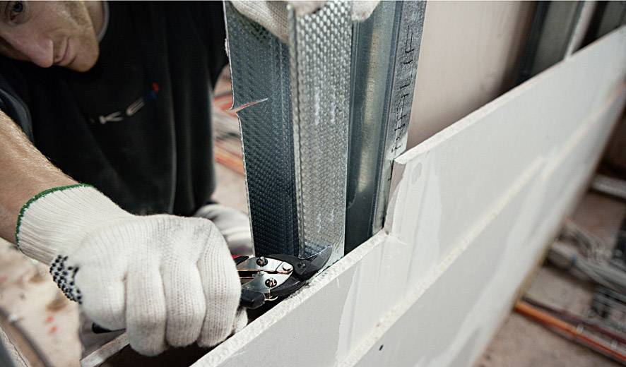 Un ouvrier fixe une construction métallique avec des pinces sur une cloison sèche. Il porte des gants de travail pour sa sécurité.