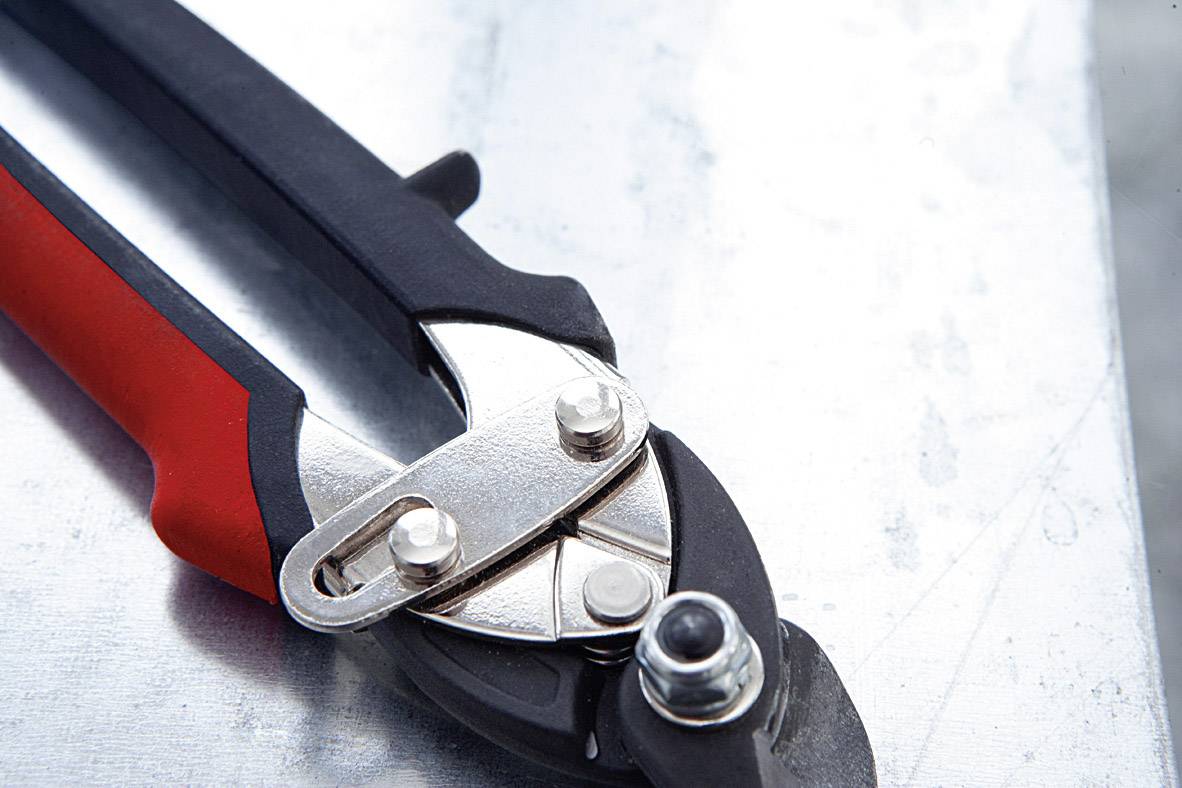 Des ciseaux de métal avec une poignée rouge et noire sont posés sur une surface métallique. Ils sont utilisés pour couper de la tôle et du fil.