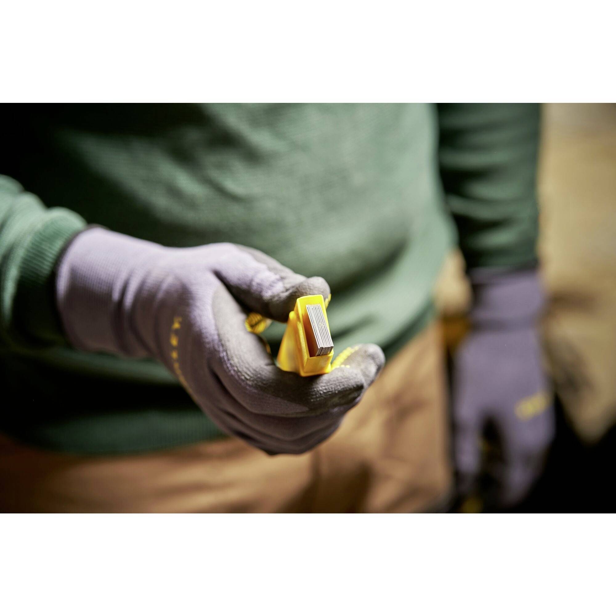 Une personne tient un outil de coupe jaune avec une lame à la main, porte des gants et un pull vert ; l'accent est mis sur l'outil.